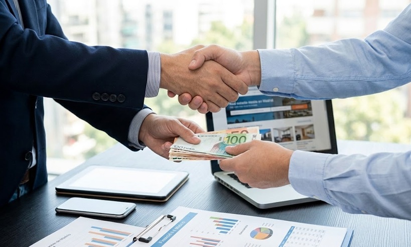 Contrato de earn out en España con apretón de manos y entrega de euros sobre escritorio con informes de beneficios y due diligence financiera.